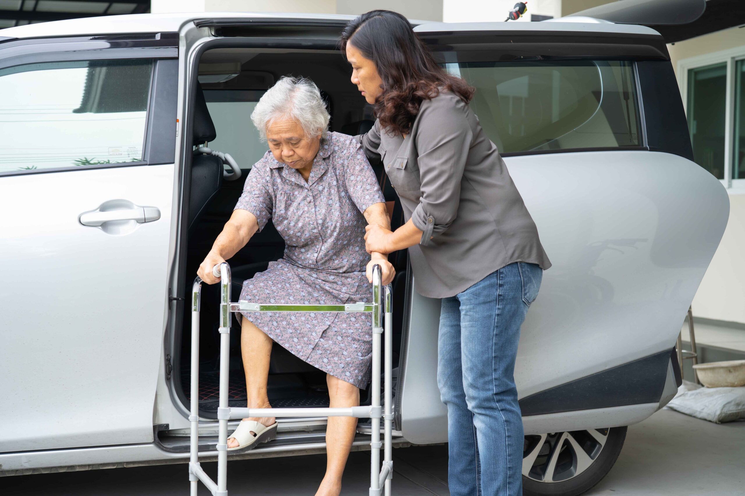 Asian senior woman sitting on a walker while preparing to stand with caregiver support.